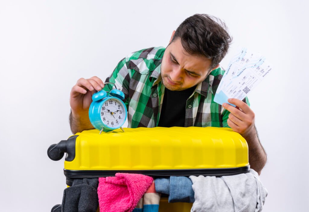 young traveler man in checked shirt standing with suitcase full of clothes holding air tickets looking at alarm clock confused and very anxious over white background